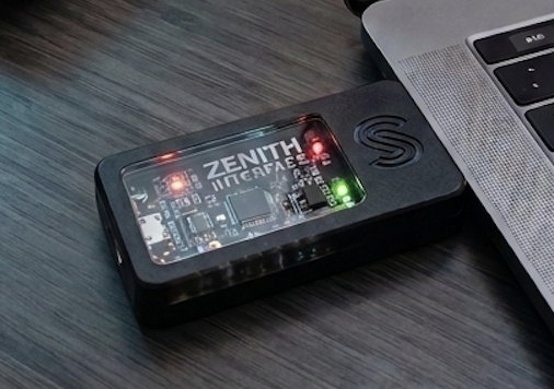 Close-up of TheClicker USB bridge plugged into a MacBook, showing the Zenith Interface circuit board with red and green status LEDs
